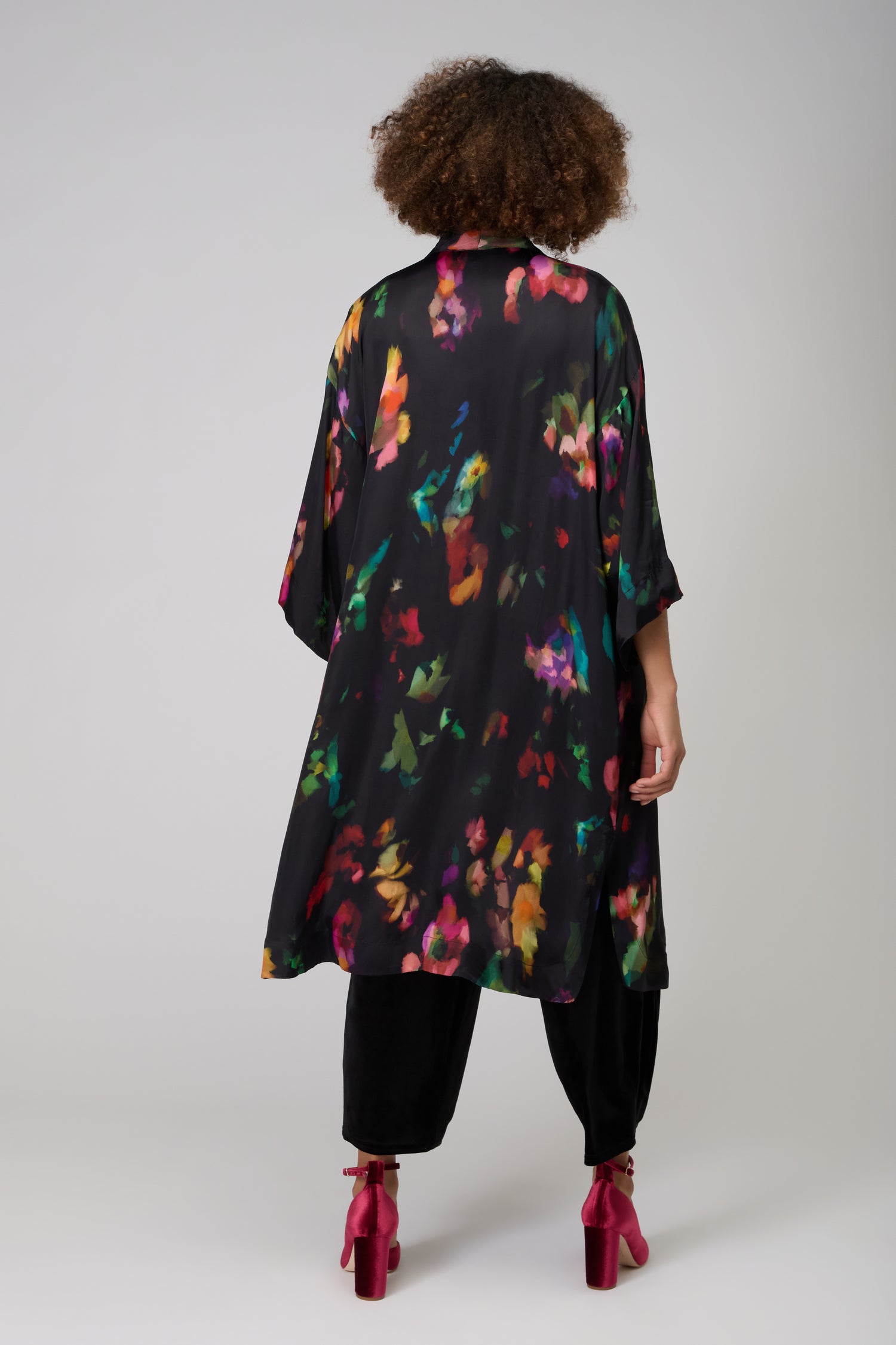 A person stands with their back to the camera in a Winter Floral Kimono, featuring a painterly floral print, styled with black pants and red heels against a plain studio background.