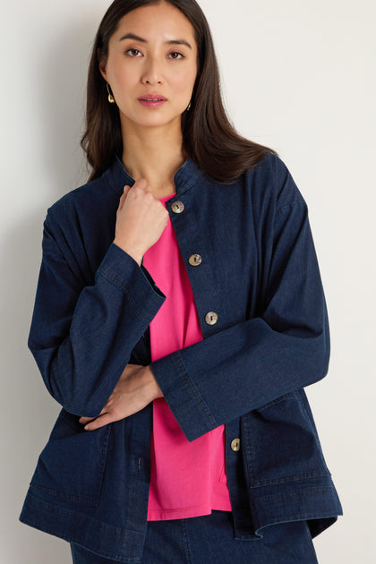 A woman wears the Stretch Denim Boxy Jacket, a relaxed-fit dark stretch denim layer, over a bright pink top, standing against a plain light-colored background.