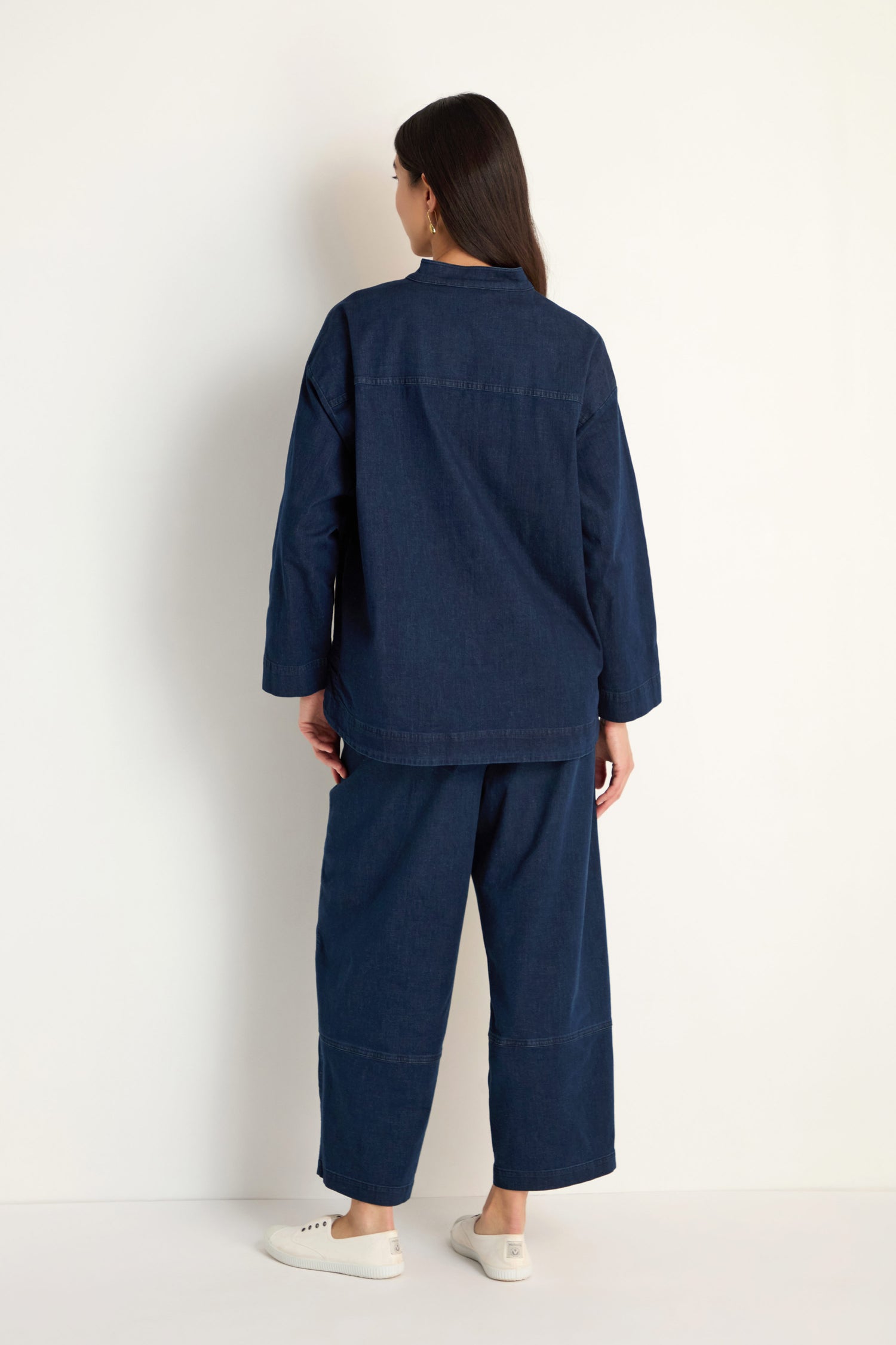 A person stands facing away, wearing the Stretch Denim Boxy Jacket in dark blue with matching wide-leg pants and white sneakers, set against a plain light background.