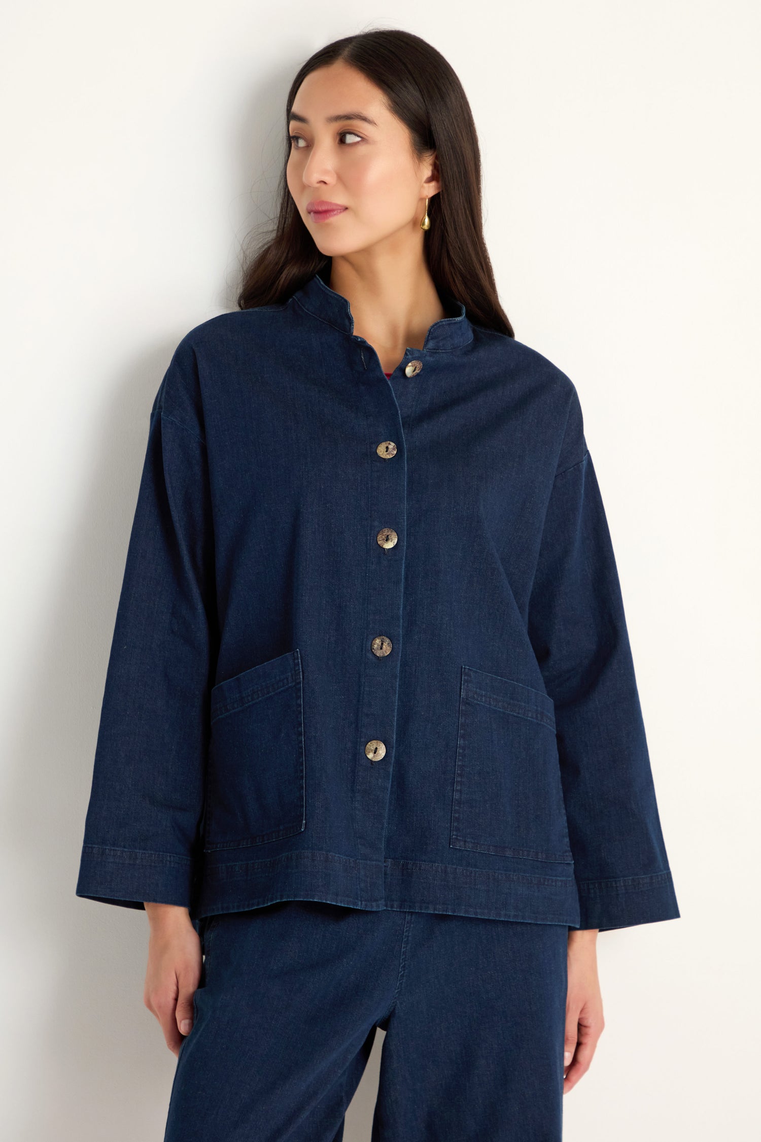 A woman wears the Stretch Denim Boxy Jacket in dark blue with large buttons and front pockets, paired with matching denim pants, as she stands against a plain white background.