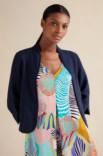 A woman with dark hair in a low ponytail wears the Navy Linen Cropped Jacket over a colorful, patterned dress, standing against a neutral background.