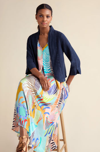 A woman sits on a wooden stool wearing a Navy Linen Cropped Jacket layered over a colorful patterned maxi dress, against a neutral background.