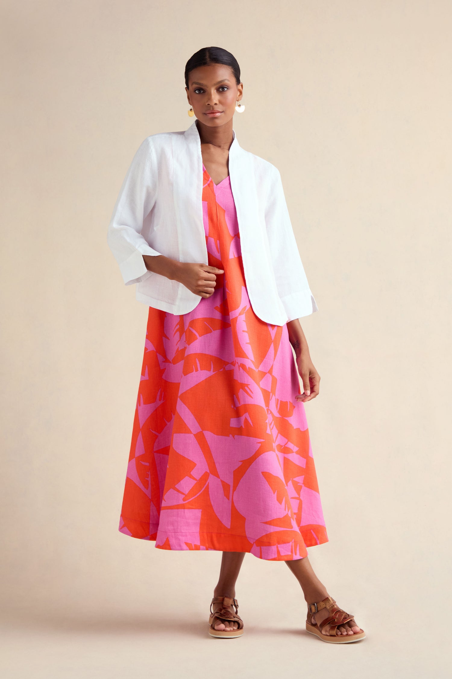 A woman stands against a plain beige background wearing a White Linen Cropped Jacket over a pink and orange patterned midi dress with brown sandals.