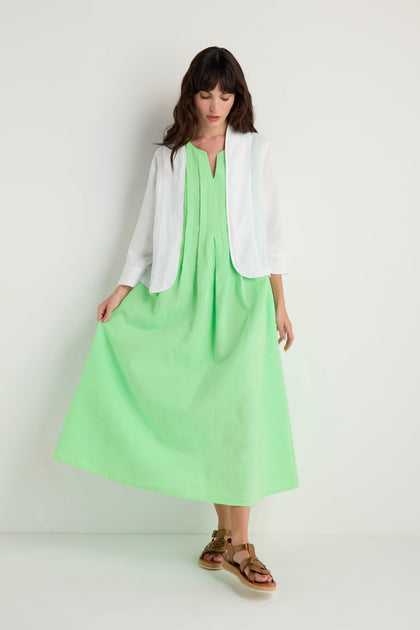 A woman stands against a plain white background, wearing a boxy fit White Linen Cropped Jacket over a mint green ankle-length dress with brown sandals.