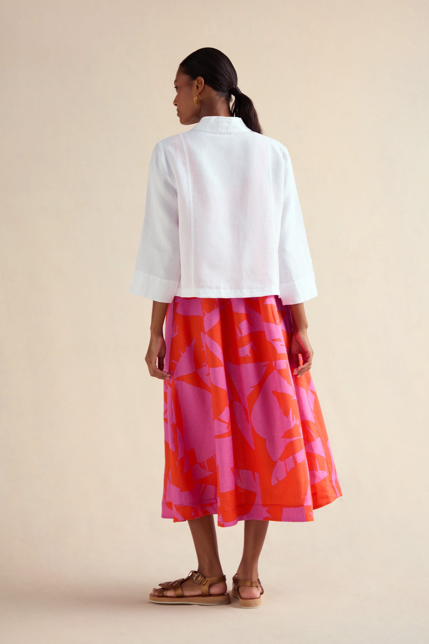 A woman, seen from behind against a beige background, wears the White Linen Cropped Jacket layered over a pink and red patterned midi skirt, paired with tan sandals and gold earrings.