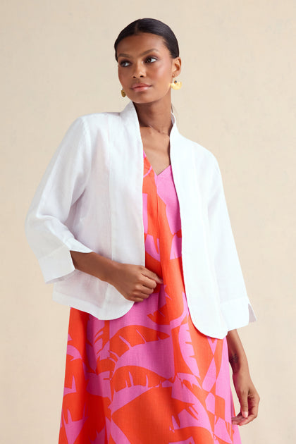 Model wears the White Linen Cropped Jacket as a chic layer over a bold pink and orange patterned dress, standing against a neutral background.