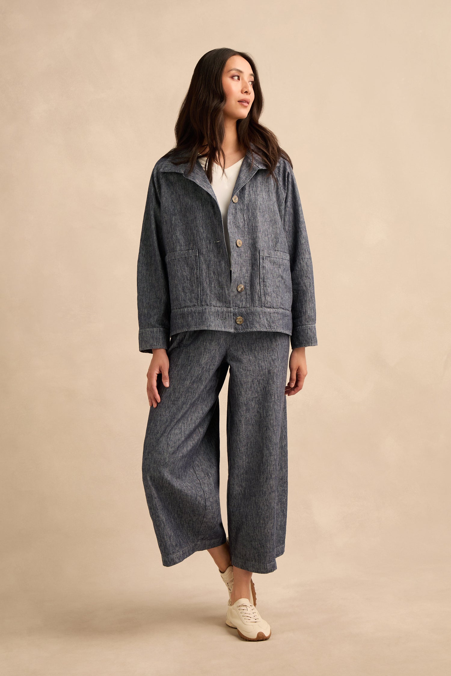 A woman stands against a plain background, wearing a loose-fitting Washed Cotton Linen Jacket and wide-leg pants with white sneakers, looking slightly to the side.