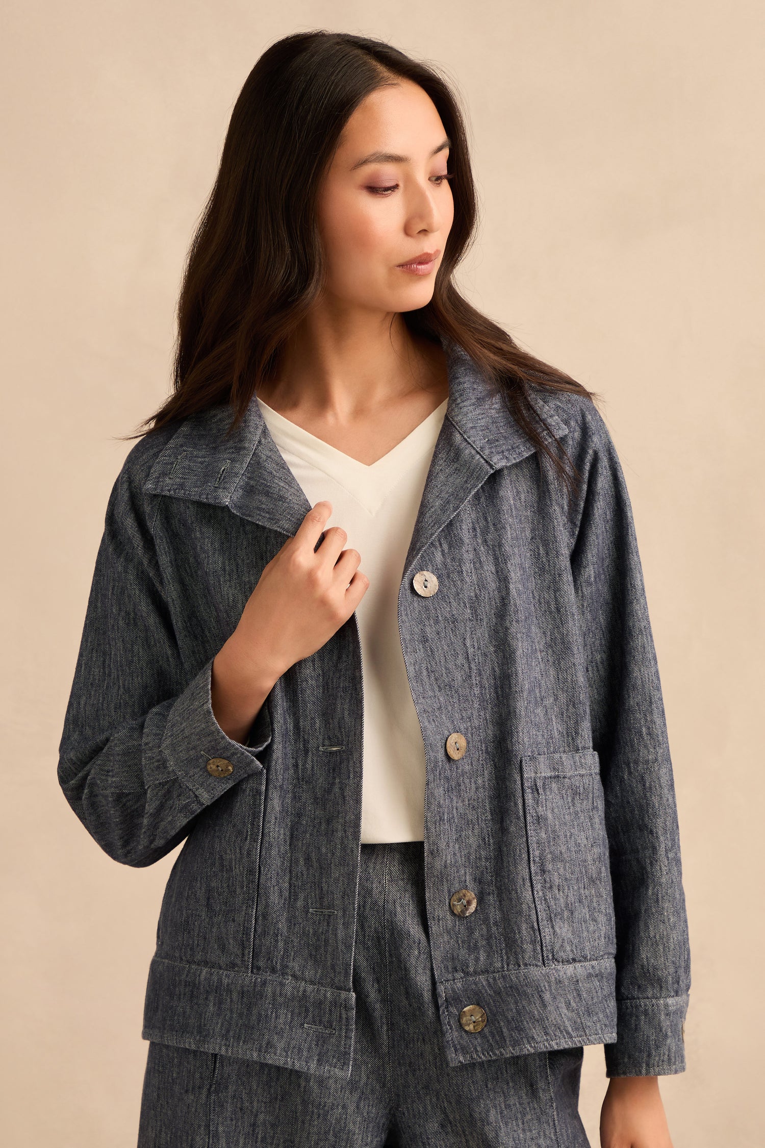 A woman in a Washed Cotton Linen Jacket and matching pants stands against a beige background, looking to her left with her hand touching the jacket.