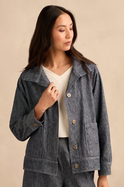 A woman in a Washed Cotton Linen Jacket and matching pants stands against a beige background, looking to her left with her hand touching the jacket.