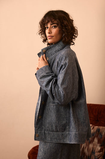 A person with curly hair stands sideways, looking at the camera, wearing the Washed Cotton Linen Jacket buttoned up with matching pants. The scene has a neutral indoor background and a partial view of a chair.