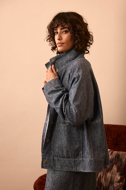 A person with curly hair stands sideways, looking at the camera, wearing the Washed Cotton Linen Jacket buttoned up with matching pants. The scene has a neutral indoor background and a partial view of a chair.