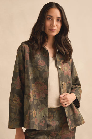 A woman stands against a plain beige background, looking at the camera, wearing the Heirloom Damask Denim Jacket featuring a vintage archive print over a cream top.