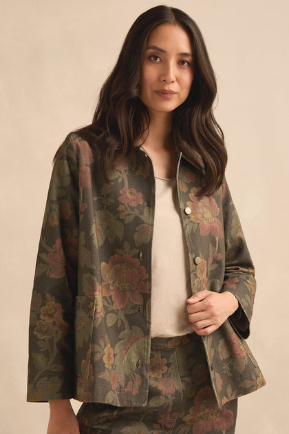 A woman stands against a plain beige background, looking at the camera, wearing the Heirloom Damask Denim Jacket featuring a vintage archive print over a cream top.