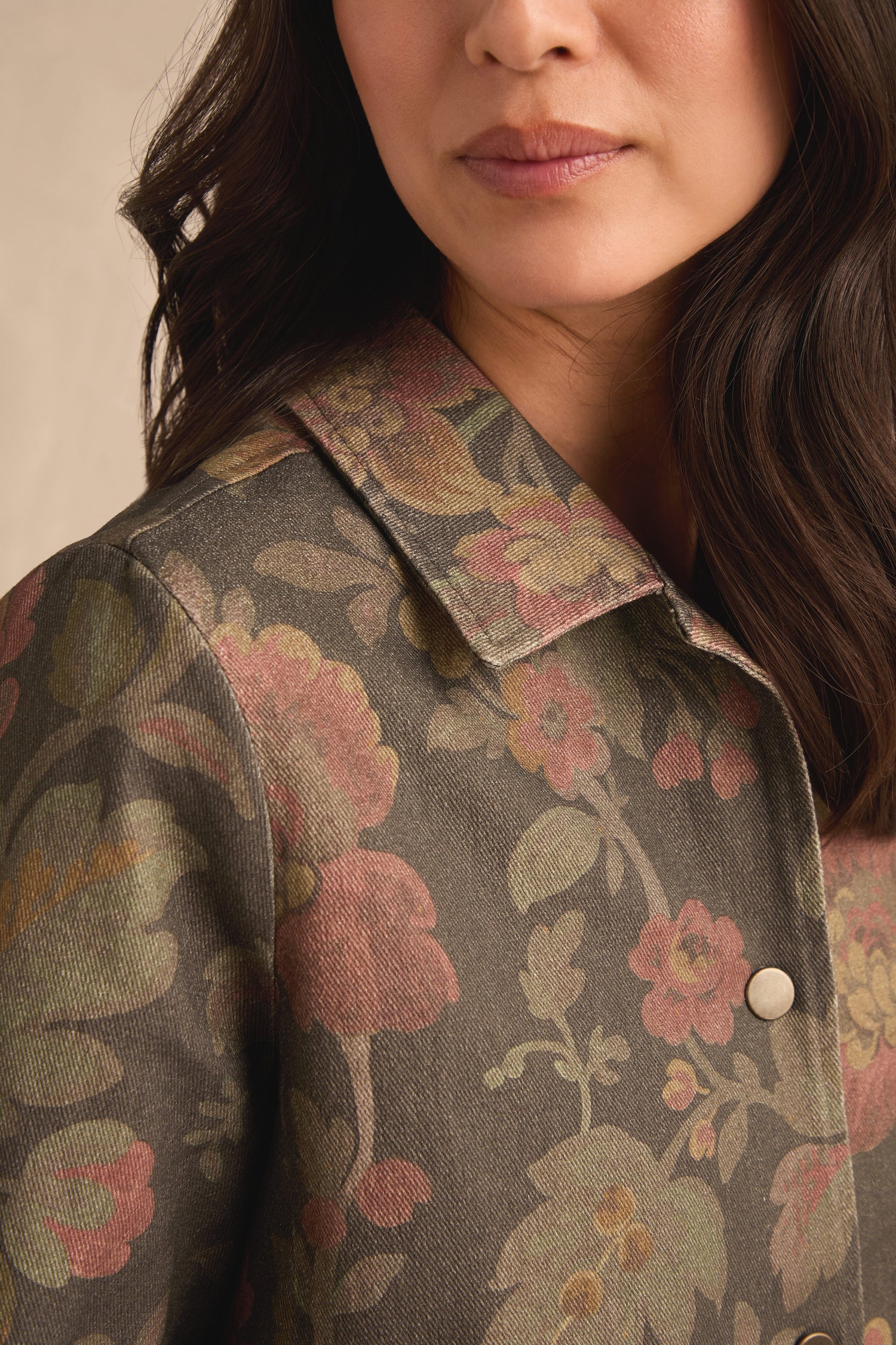 A woman models the Heirloom Damask Denim Jacket—a pure cotton, collared piece featuring a muted floral pattern in green, pink, and yellow tones inspired by vintage archive prints. Only the lower half of her face and upper torso are visible.