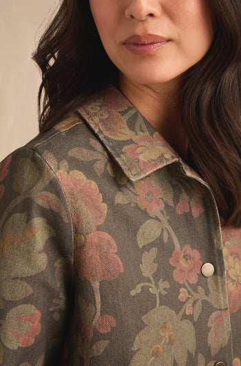 A woman models the Heirloom Damask Denim Jacket—a pure cotton, collared piece featuring a muted floral pattern in green, pink, and yellow tones inspired by vintage archive prints. Only the lower half of her face and upper torso are visible.