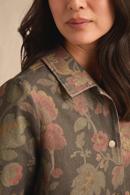 A woman models the Heirloom Damask Denim Jacket—a pure cotton, collared piece featuring a muted floral pattern in green, pink, and yellow tones inspired by vintage archive prints. Only the lower half of her face and upper torso are visible.