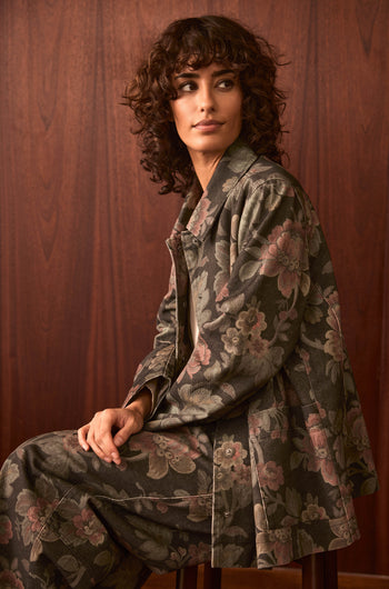 A person with curly brown hair sits sideways on a chair, wearing the Heirloom Damask Denim Jacket featuring a vintage archive floral print, paired with matching pants, in front of a wooden wall.