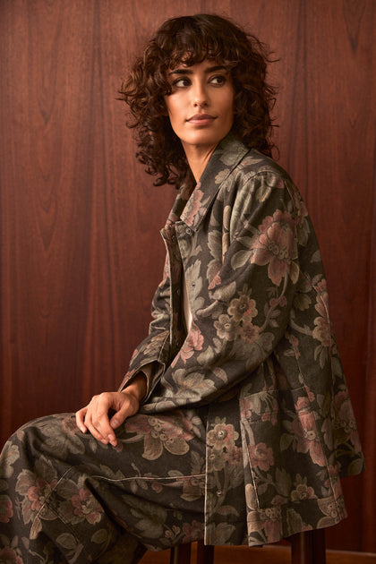 A person with curly brown hair sits sideways on a chair, wearing the Heirloom Damask Denim Jacket featuring a vintage archive floral print, paired with matching pants, in front of a wooden wall.