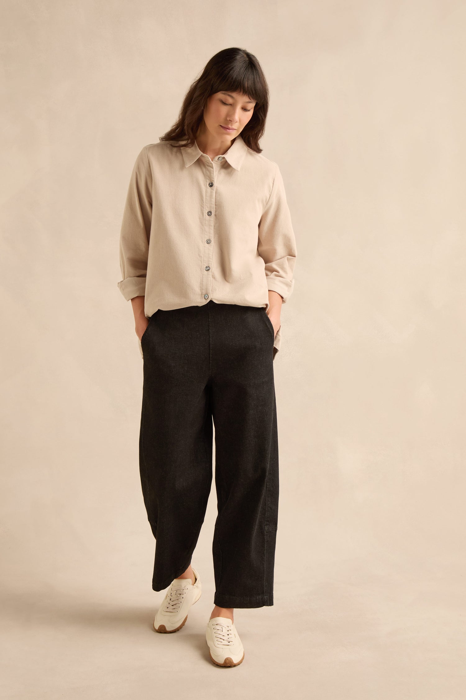 Woman wearing a beige button-up shirt, Stretch Black Denim Bubble Trousers, and white sneakers stands with hands in pockets against a plain beige background.