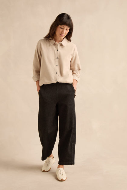Woman wearing a beige button-up shirt, Stretch Black Denim Bubble Trousers, and white sneakers stands with hands in pockets against a plain beige background.