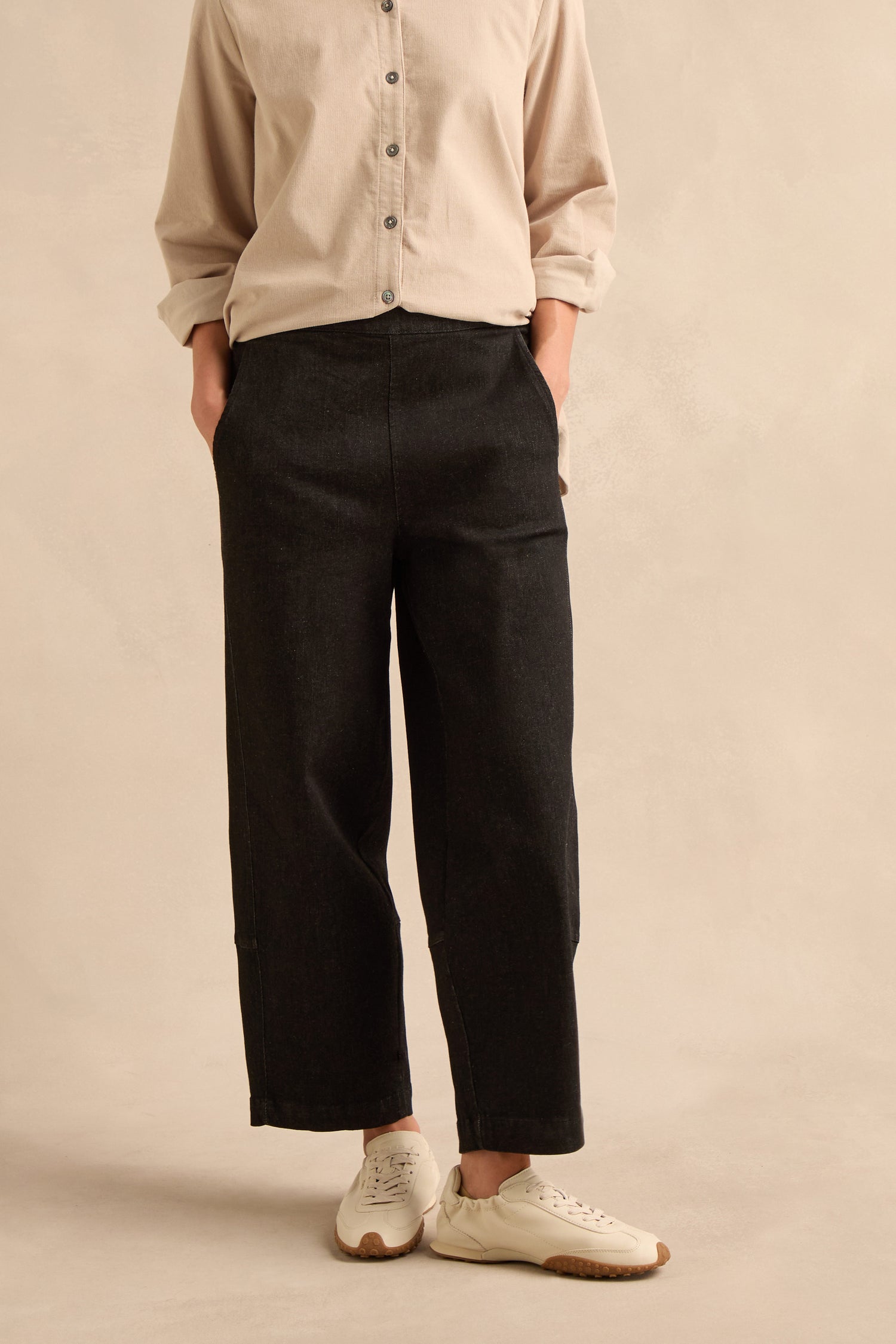 A person stands with hands in pockets against a neutral background, wearing a beige button-up shirt, Stretch Black Denim Bubble Trousers with a cropped leg, and white sneakers.