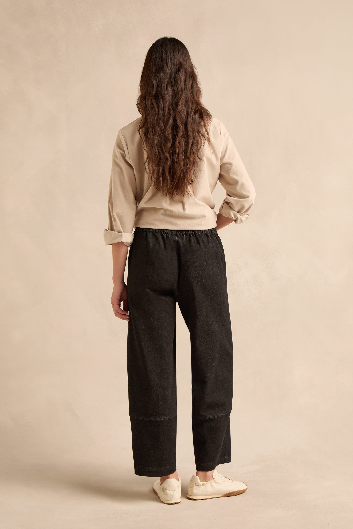 A person stands with their back to the camera, wearing beige shirt and Stretch Black Denim Bubble Trousers with a cropped leg, paired with white sneakers, against a plain beige background.