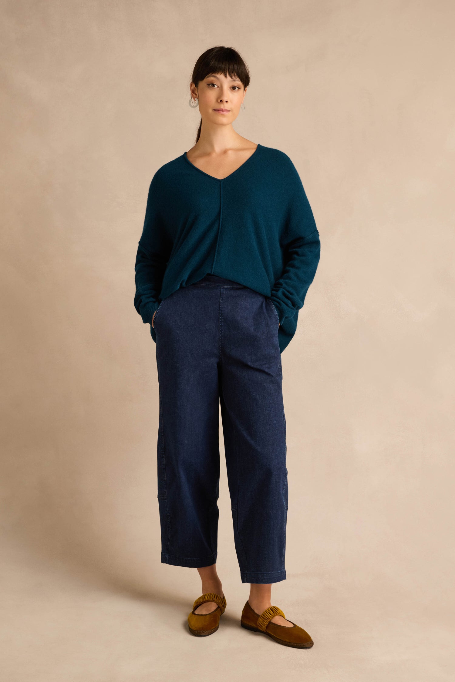 A person stands against a beige background wearing a teal sweater, the Stretch Denim Blue Bubble Trousers with an elasticated waist, and brown shoes, hands tucked in pockets.