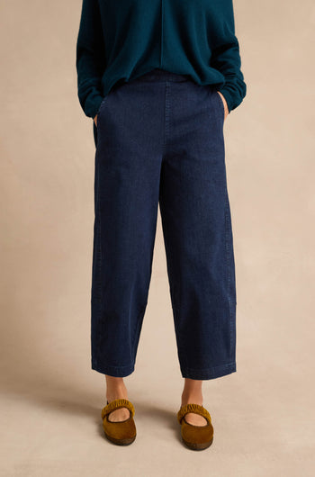 Wearing the Stretch Denim Blue Bubble Trousers—dark blue, wide-leg, cropped denim pants with an elasticated waist—paired with a dark green long-sleeve top and brown slip-on shoes, a person stands hands in pockets against a plain beige background.