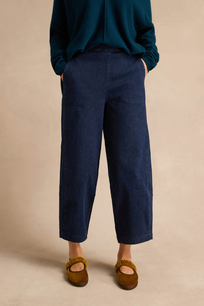 Wearing the Stretch Denim Blue Bubble Trousers—dark blue, wide-leg, cropped denim pants with an elasticated waist—paired with a dark green long-sleeve top and brown slip-on shoes, a person stands hands in pockets against a plain beige background.