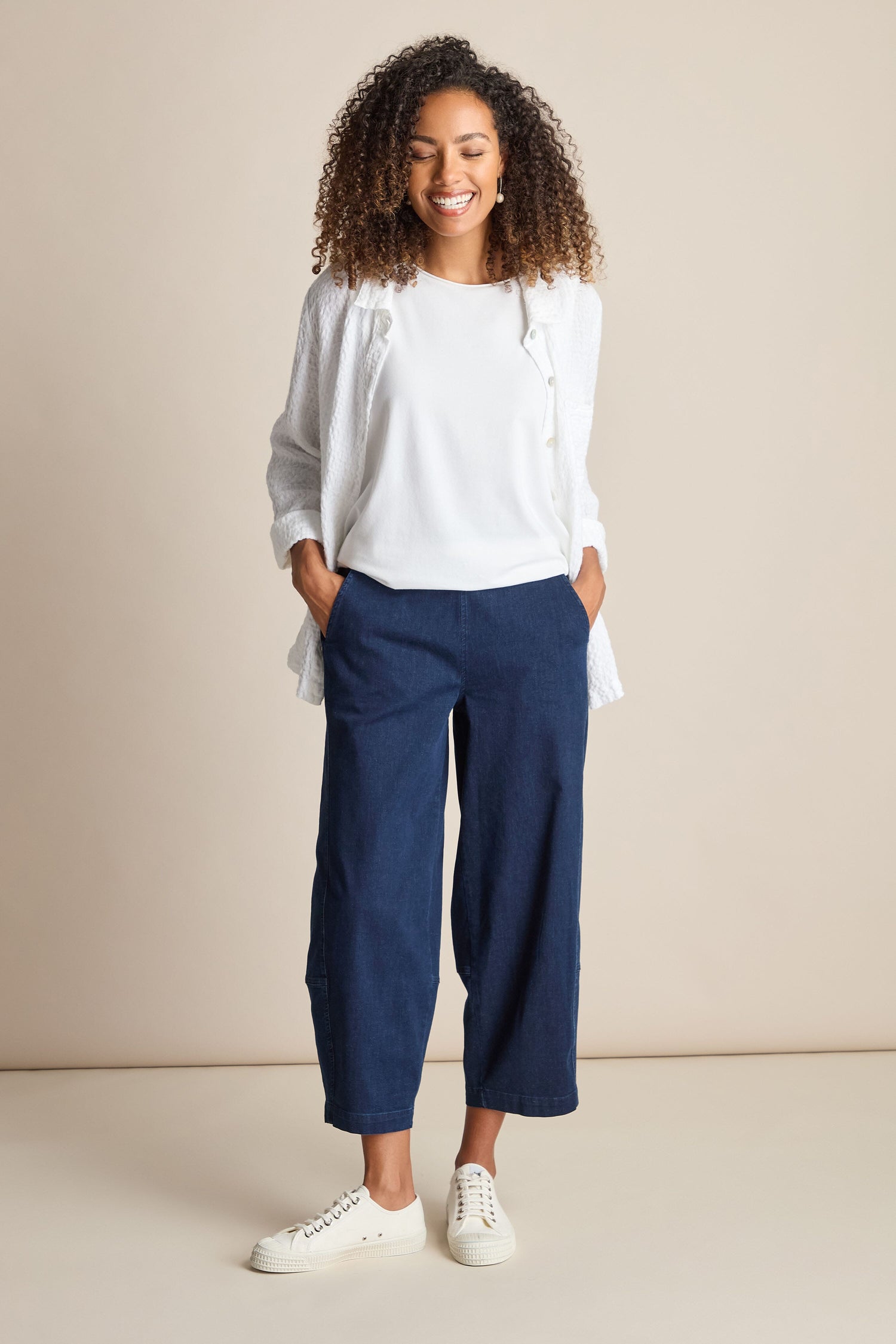 A woman stands against a neutral background, wearing a white blouse, white cardigan, the Stretch Denim Bubble Trouser in blue, and white sneakers. She has her hands in her pockets and is smiling.