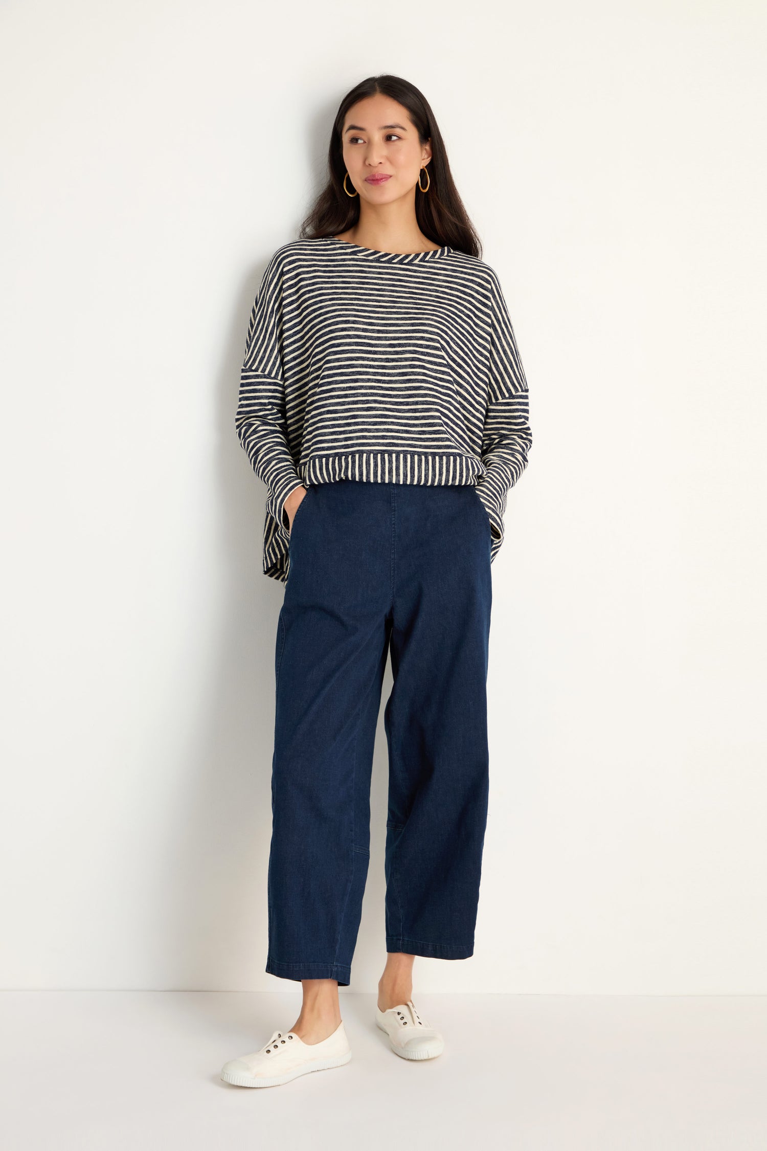 A woman stands against a plain wall wearing a black and white striped long-sleeve top, Stretch Denim Bubble Trouser in blue, and white sneakers.