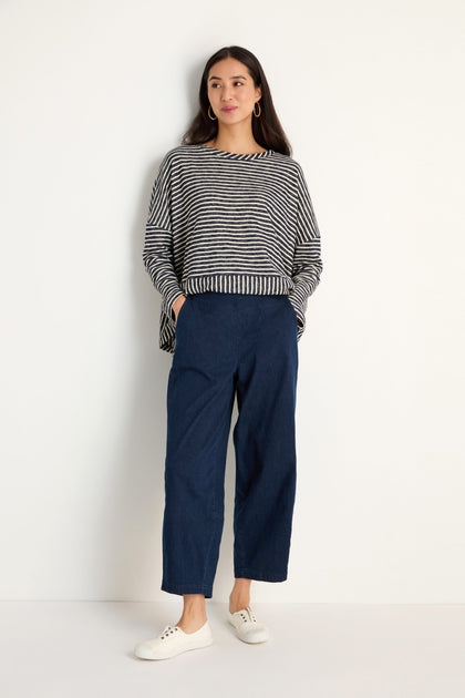 A woman stands against a plain wall wearing a black and white striped long-sleeve top, Stretch Denim Bubble Trouser in blue, and white sneakers.