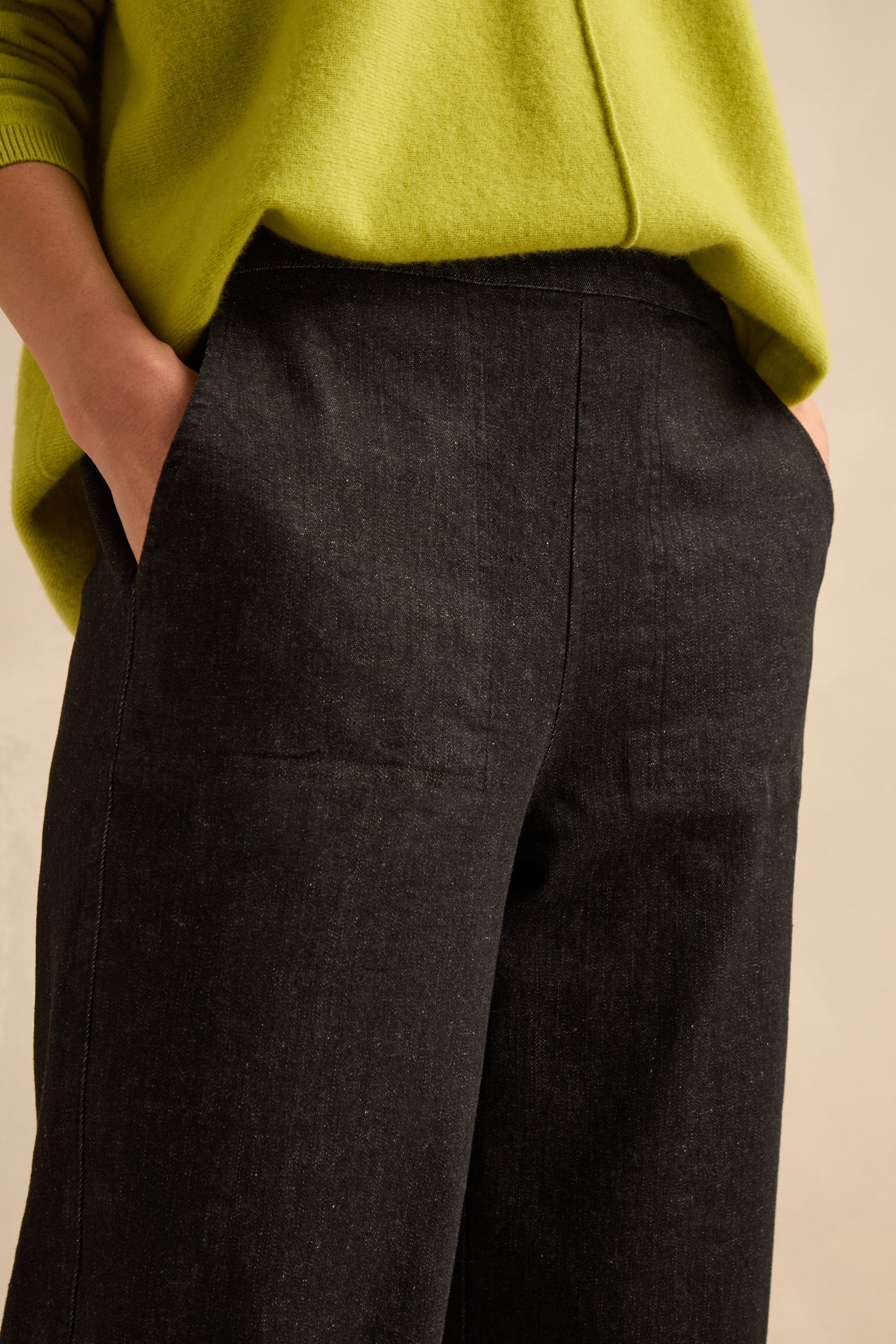 Person wearing a lime green sweater with hands in the pockets of Stretch Black Denim Straight trousers, which feature a high-waisted, elasticated waist design. Only the torso and upper legs are visible.