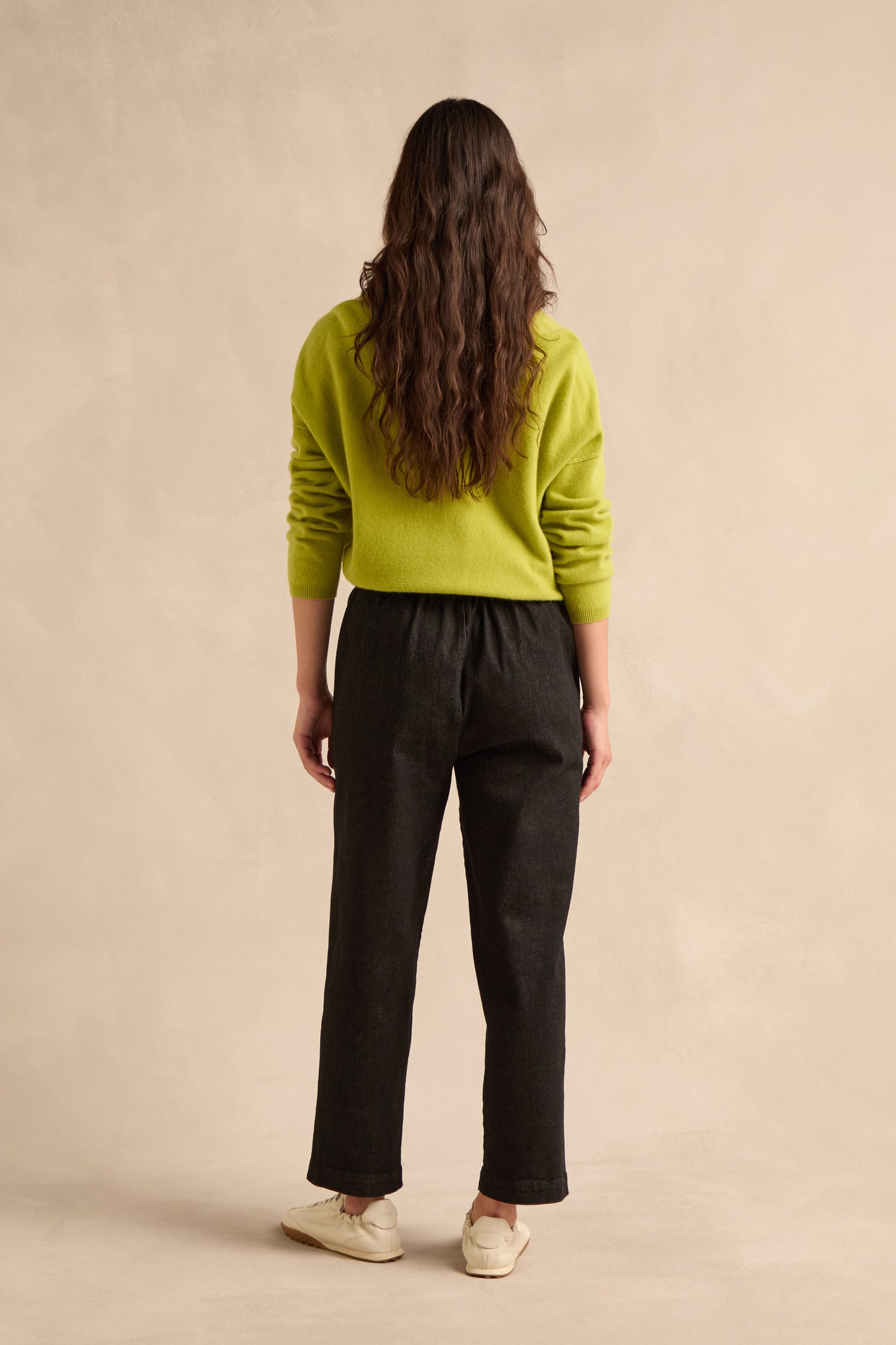 A person with long brown hair, wearing a bright green sweater, Stretch Black Denim Straight trousers with an elasticated waist, and white shoes stands facing away against a beige background.
