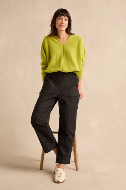 A person wearing a lime green sweater, Stretch Black Denim Straight trousers with an elasticated waist, and white sneakers stands with one leg on a wooden stool against a beige background.