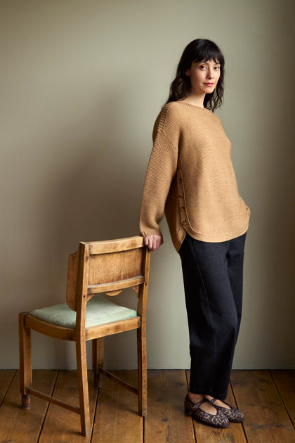 A woman wearing a tan knit sweater and Stretch Black Denim Straight Trousers stands beside a wooden chair.