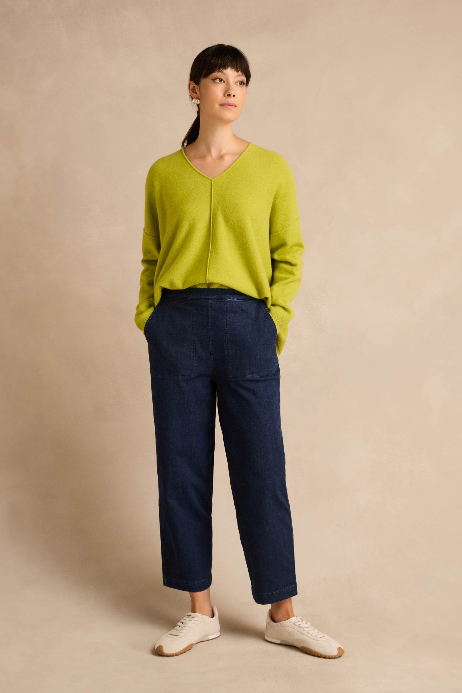 A woman stands against a neutral background, wearing a lime green sweater, the Stretch Denim Blue Straight Trousers in a pull-on style, and white sneakers, with her hands in her pockets and looking slightly to the side.