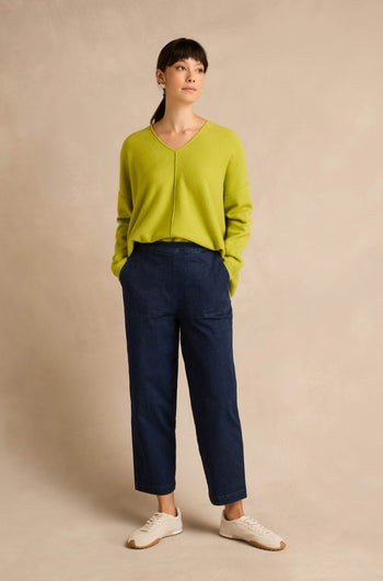A woman stands against a neutral background, wearing a lime green sweater, the Stretch Denim Blue Straight Trousers in a pull-on style, and white sneakers, with her hands in her pockets and looking slightly to the side.