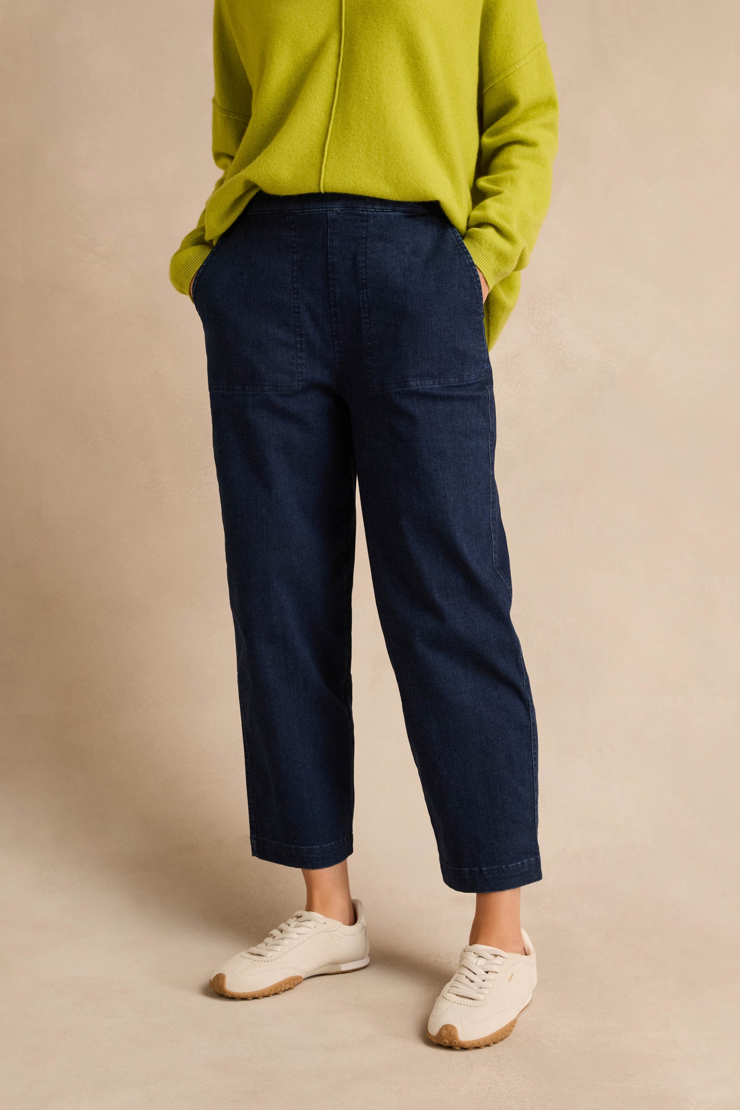 A person wears a lime green sweater, Stretch Denim Blue Straight Trousers in a pull-on style, and white sneakers, standing with hands in pockets against a beige background.