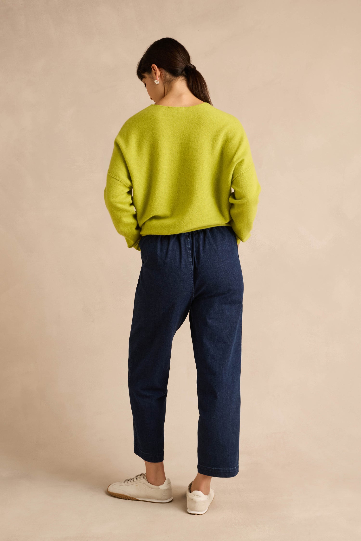 A person stands with their back to the camera, wearing a lime green sweater, Stretch Denim Blue Straight Trousers in a pull-on style, and white sneakers against a plain beige background.