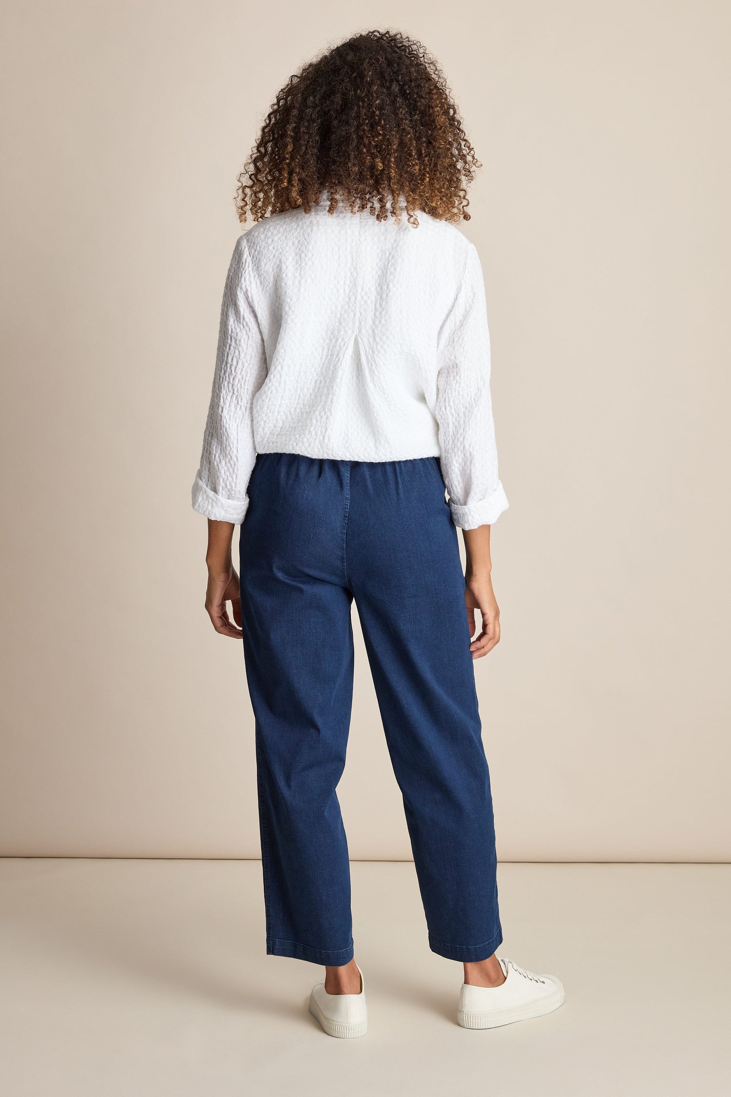 A person with curly hair is standing facing away, wearing a white shirt, Stretch Denim Straight Trouser in blue, and white sneakers against a beige backdrop.