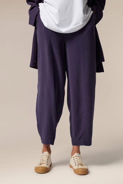 A model wears an open purple button-up shirt, white top underneath and purple wide trousers with tapered ankles. She wears neutral trainers.