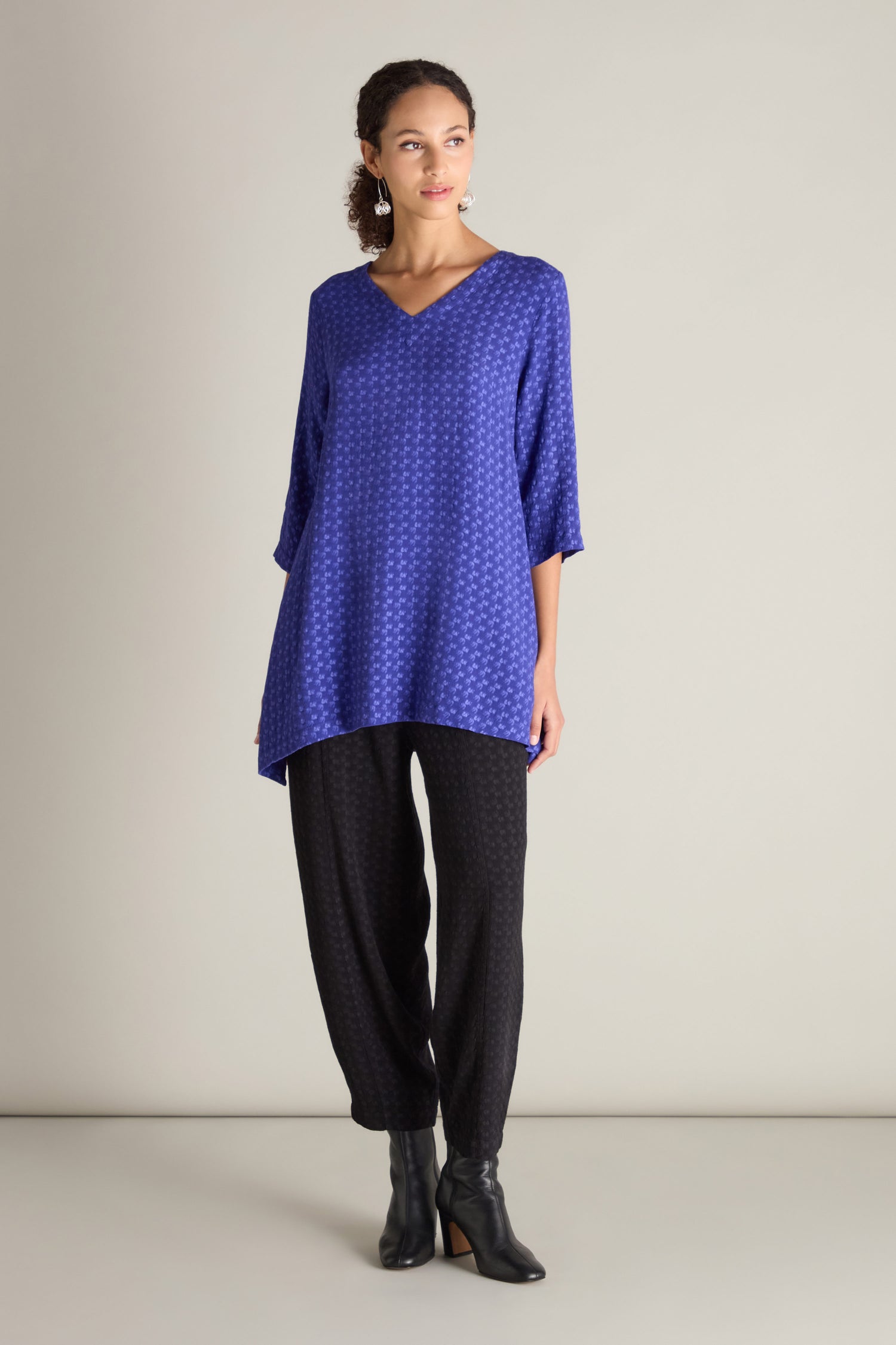 A person stands against a plain background, showcasing a chic silhouette in a blue patterned long-sleeve top paired with the Small Square Jacquard Bubble Trouser featuring an elasticated waistband and black ankle boots.