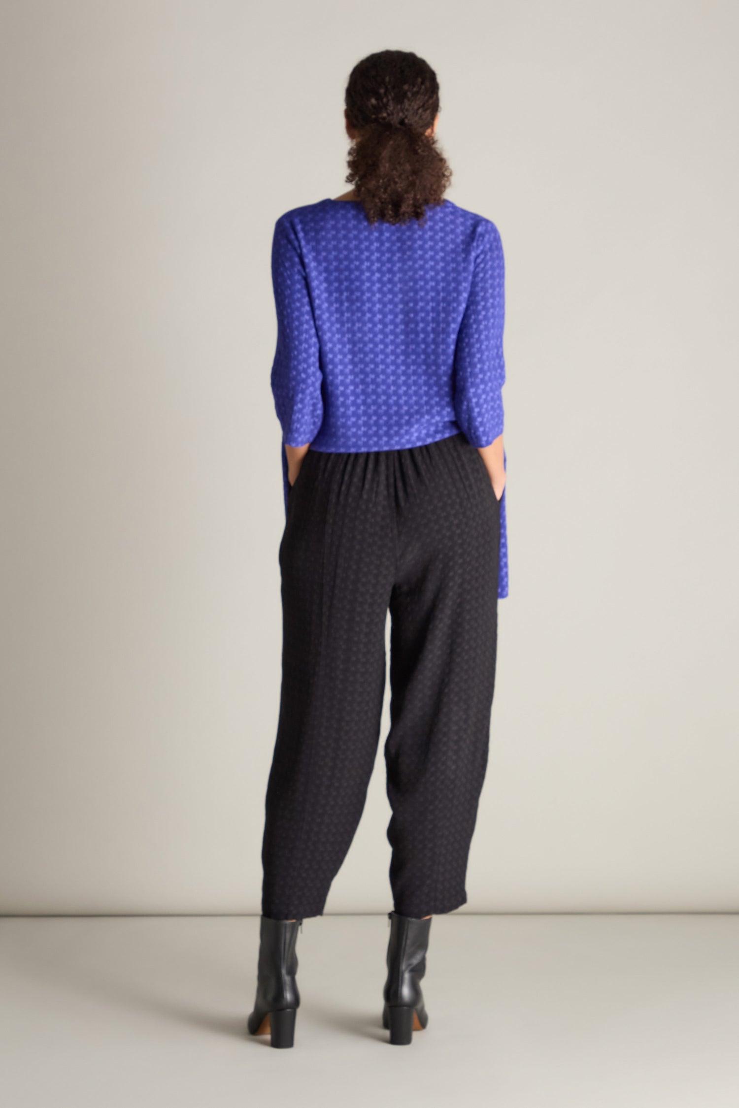 A person stands facing away, showcasing a chic silhouette in a purple top and black ankle boots, paired with Small Square Jacquard Bubble Trouser featuring an elasticated waistband.