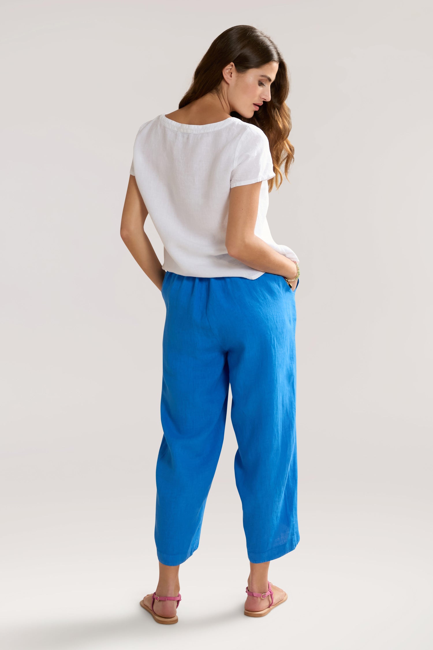 A woman with long hair is standing with her back to the camera. She is wearing a white short-sleeve top, Linen Bubble Trouser, and pink sandals.