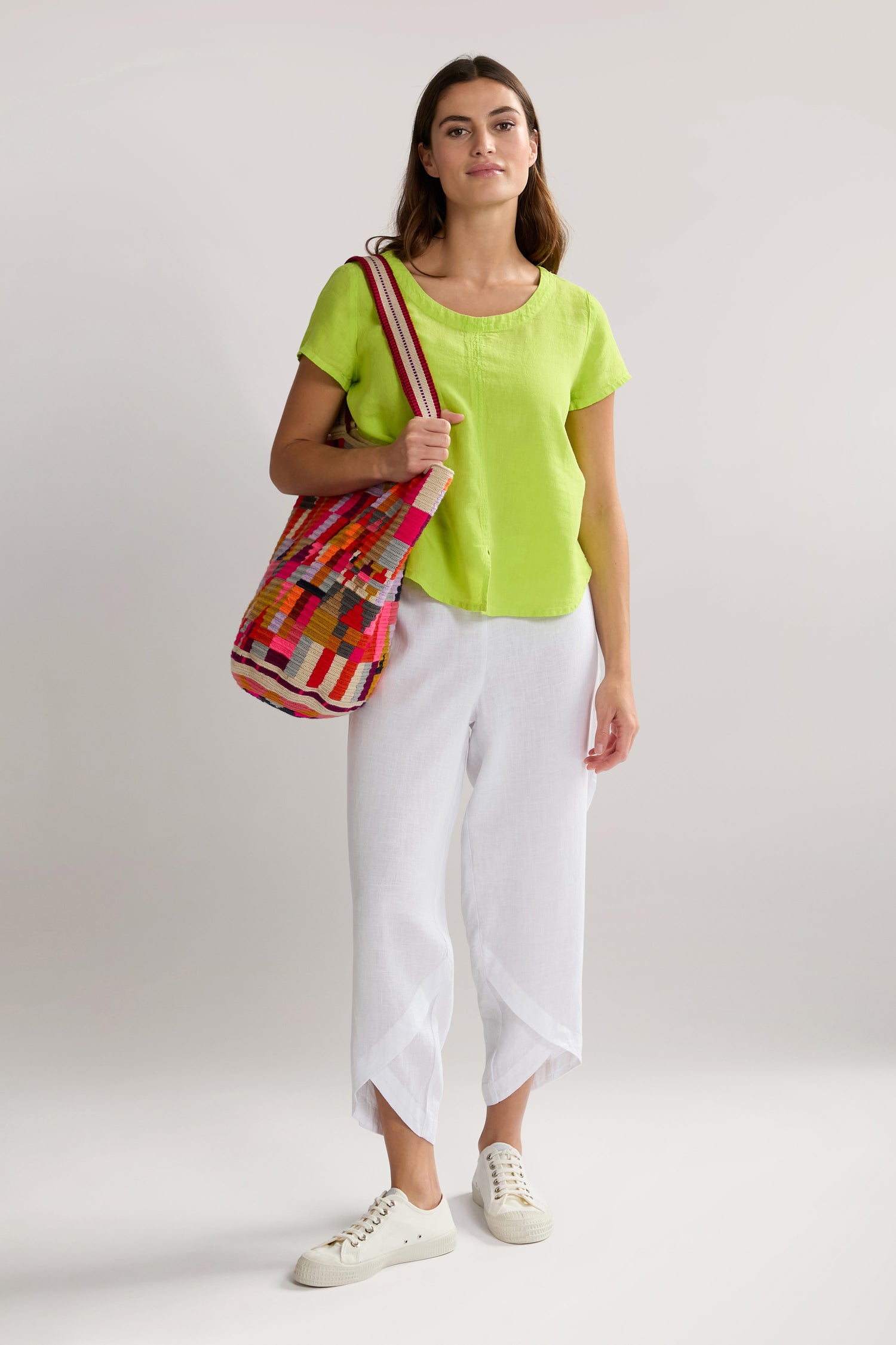 A woman stands against a plain background wearing a lime green top, Linen Bubble Trouser, white sneakers, and carrying a colorful striped bag.