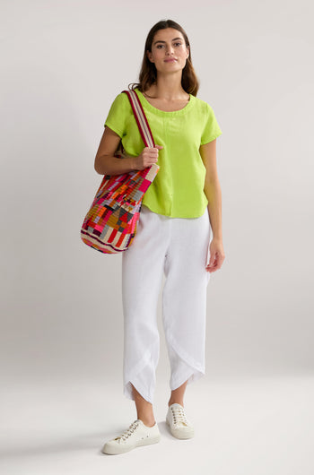 A woman stands against a plain background wearing a lime green top, Linen Bubble Trouser, white sneakers, and carrying a colorful striped bag.