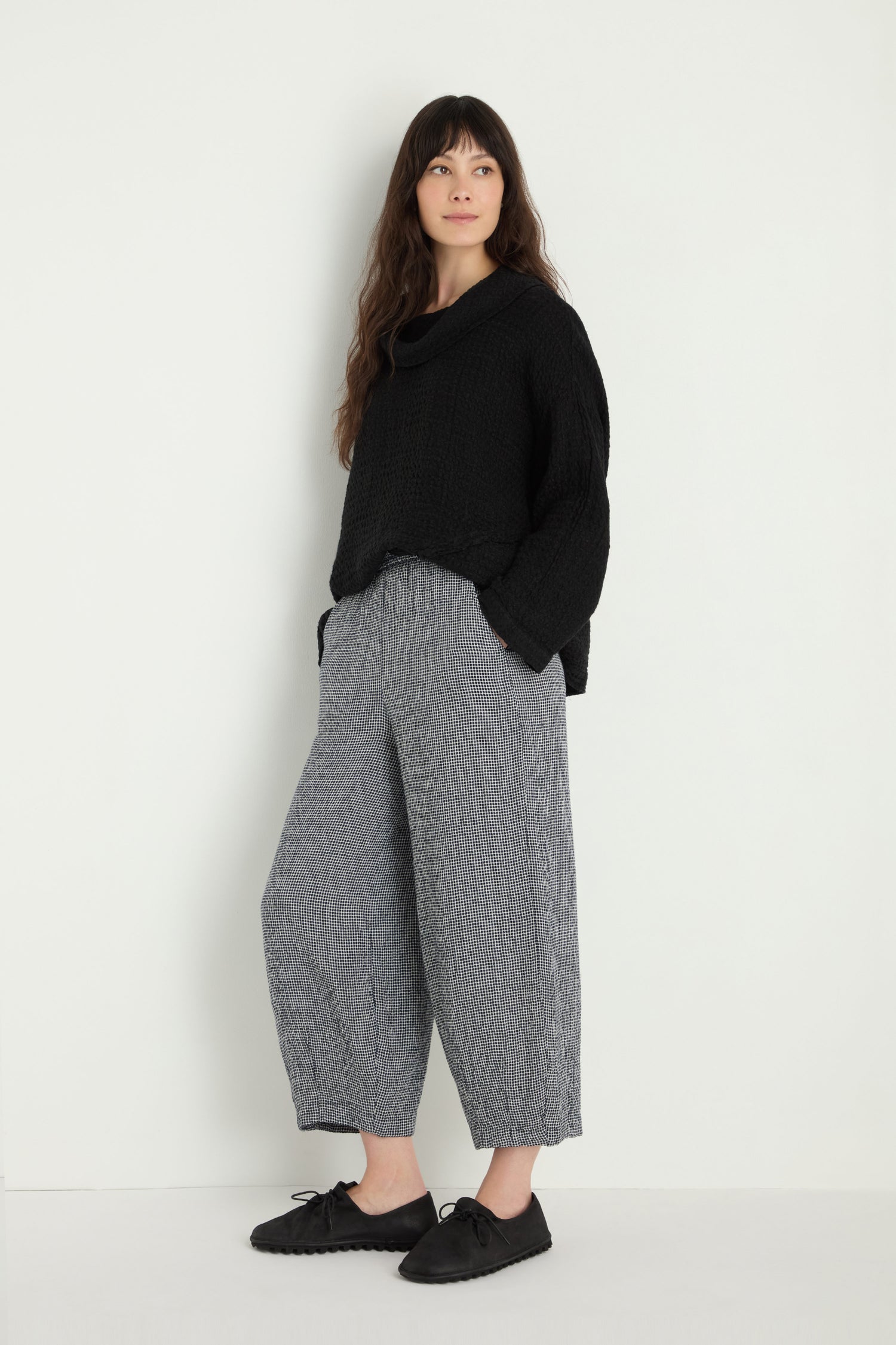 A person stands against a plain background, wearing a loose black MTP5167-MGI sweater, Micro Gingham Bubble Trouser in gray, and black shoes, with hands in pockets.