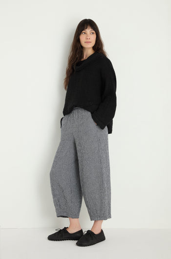 A person stands against a plain background, wearing a loose black MTP5167-MGI sweater, Micro Gingham Bubble Trouser in gray, and black shoes, with hands in pockets.