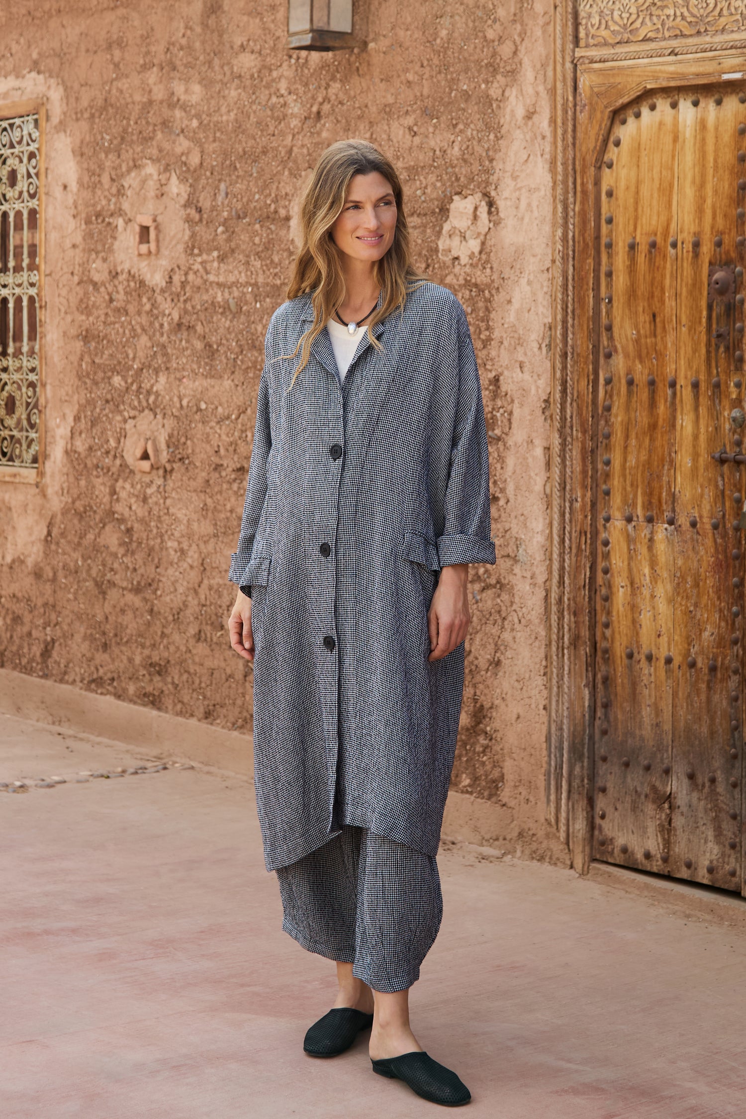 A woman stands outside by a textured wall and wooden door, wearing a loose grey coat, the Micro Gingham Bubble Trouser (MTP5167-MGI), black slippers, and a white top.